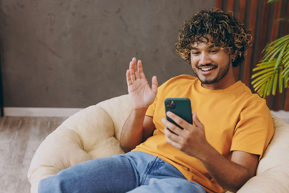 Smiling person sitting on a couch, holding a smartphone and waving during a video call in a bright living room.