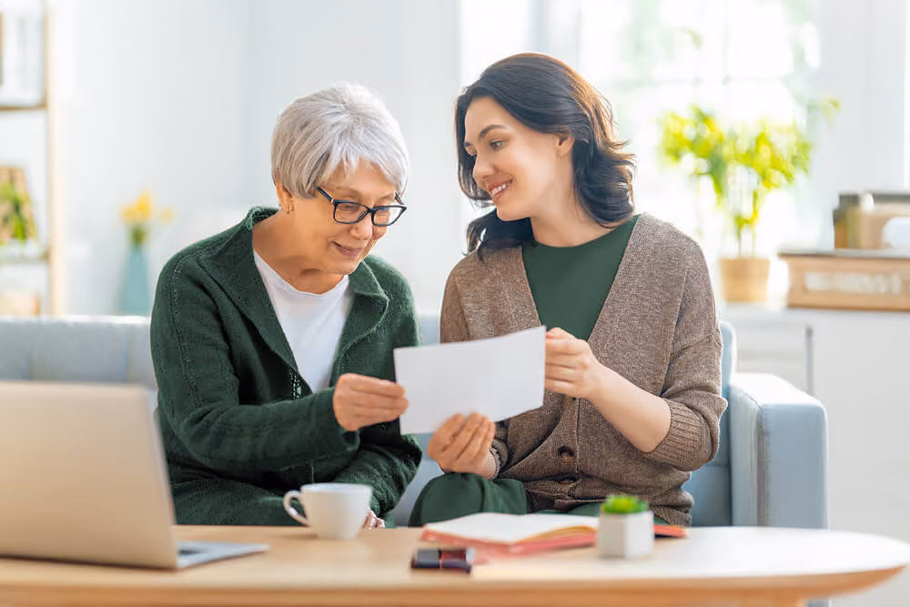 Older woman and younger woman sitting together on a couch, reviewing a document while a laptop and coffee sit on the table in a bright living room.