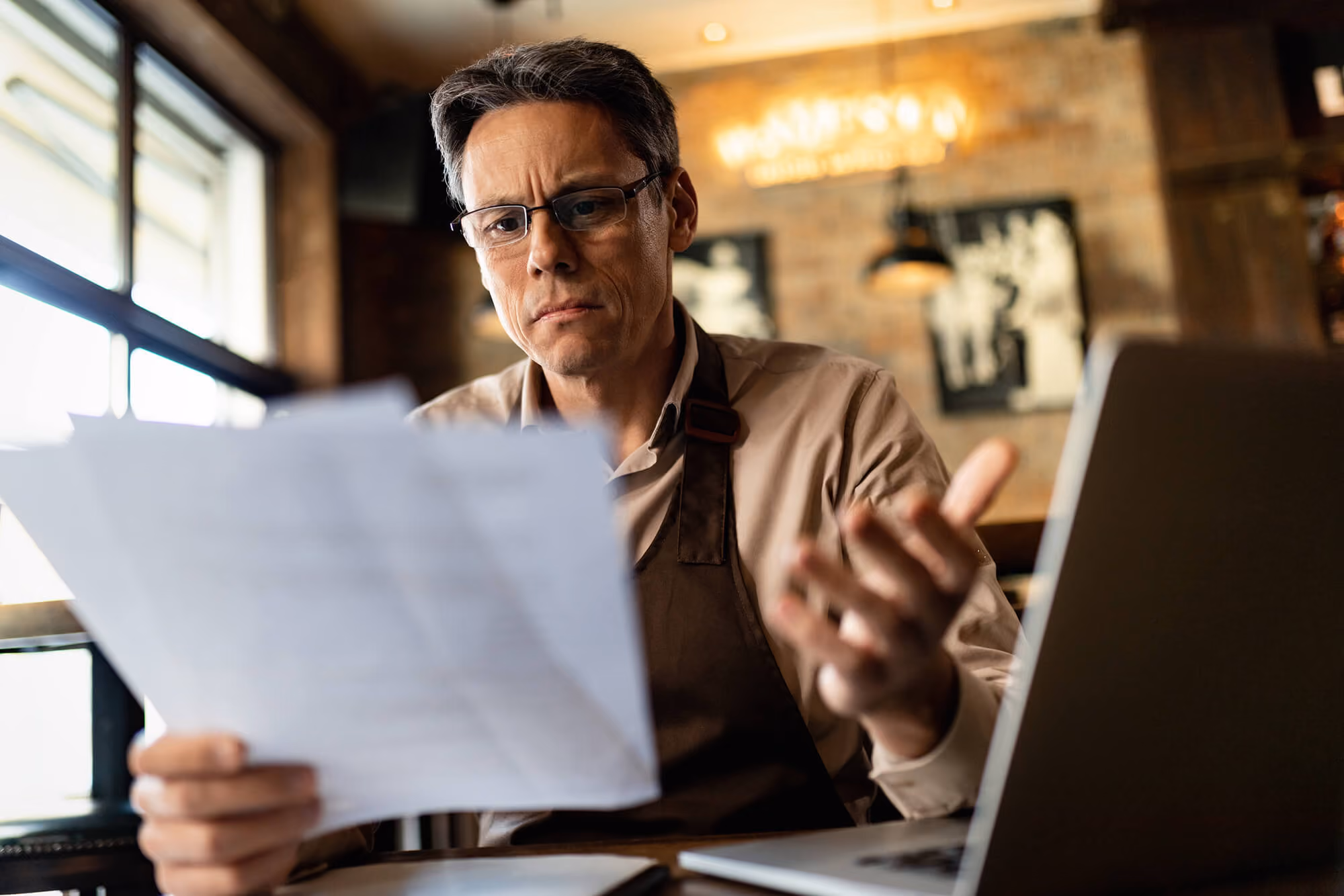 Man wearing glasses and an apron looks concerned while reviewing a document at a table with a laptop in a café setting.