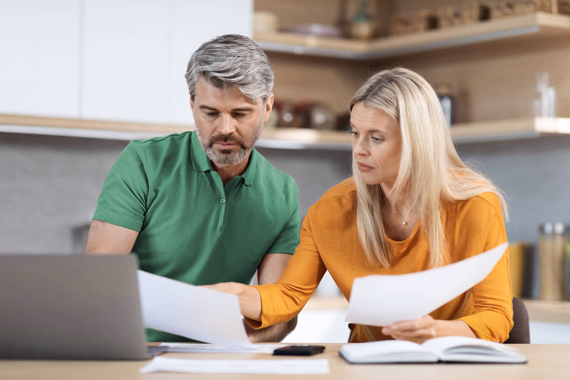 Two adults sit at a table reviewing papers together beside a laptop, looking focused as they compare documents.