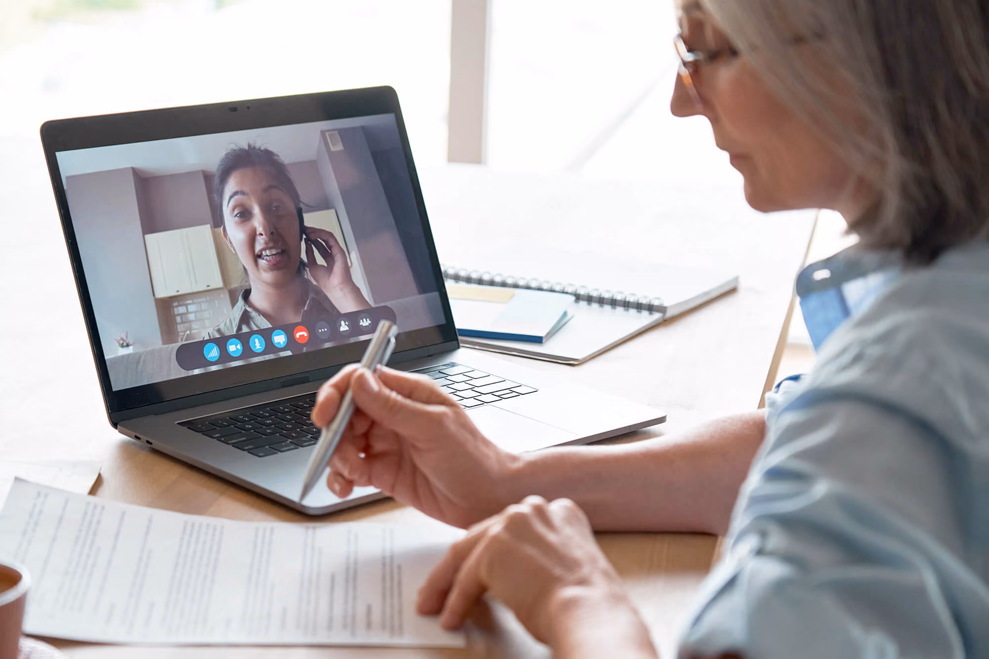 Person reviewing a document while speaking with someone on a video call displayed on a laptop screen.