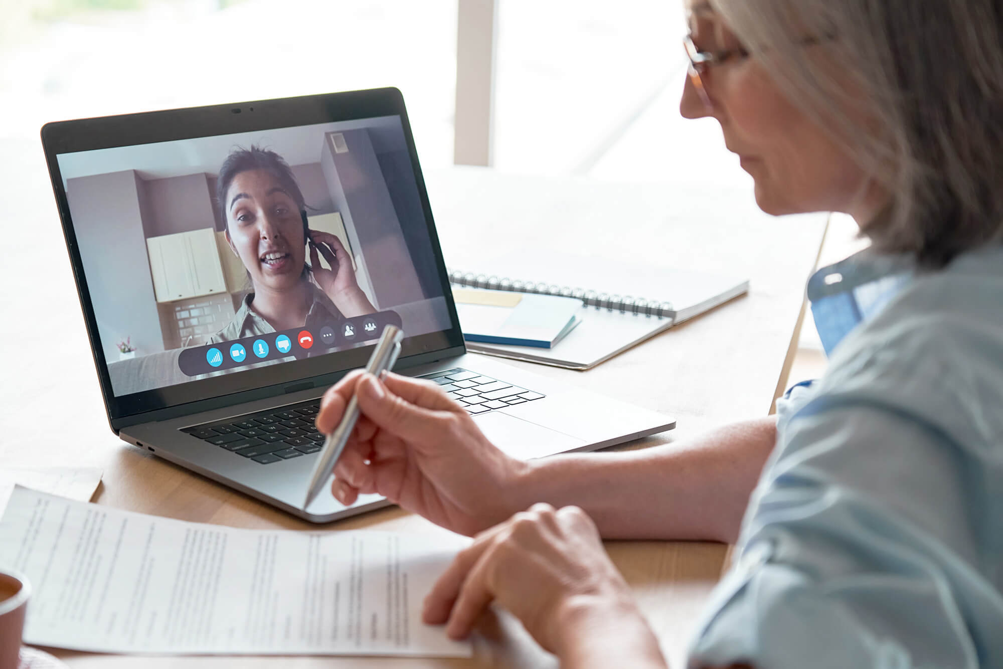 Person reviewing a document while speaking with someone on a video call displayed on a laptop screen.