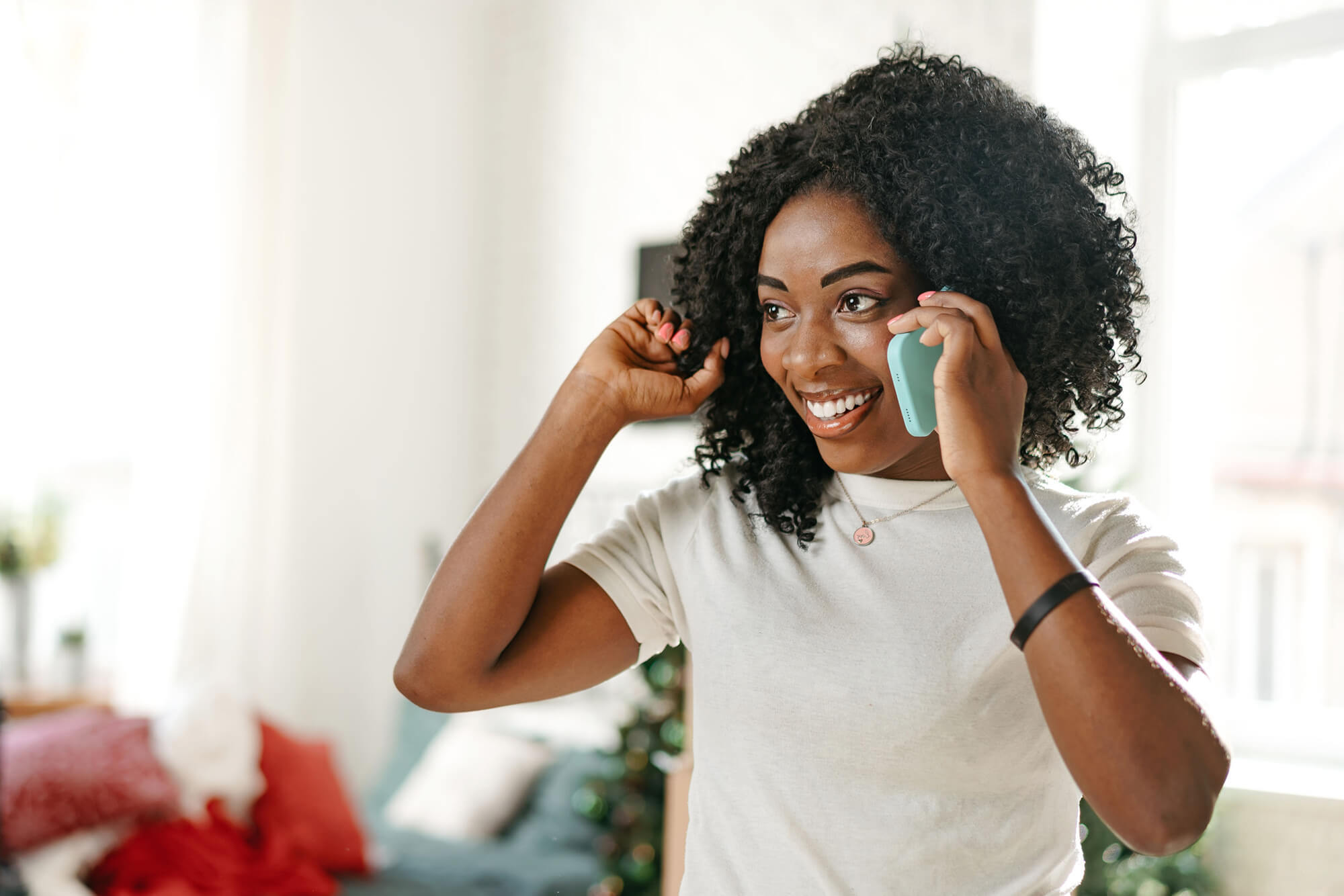 Smiling woman holding a phone to her ear while adjusting her hair, standing in a bright living room.