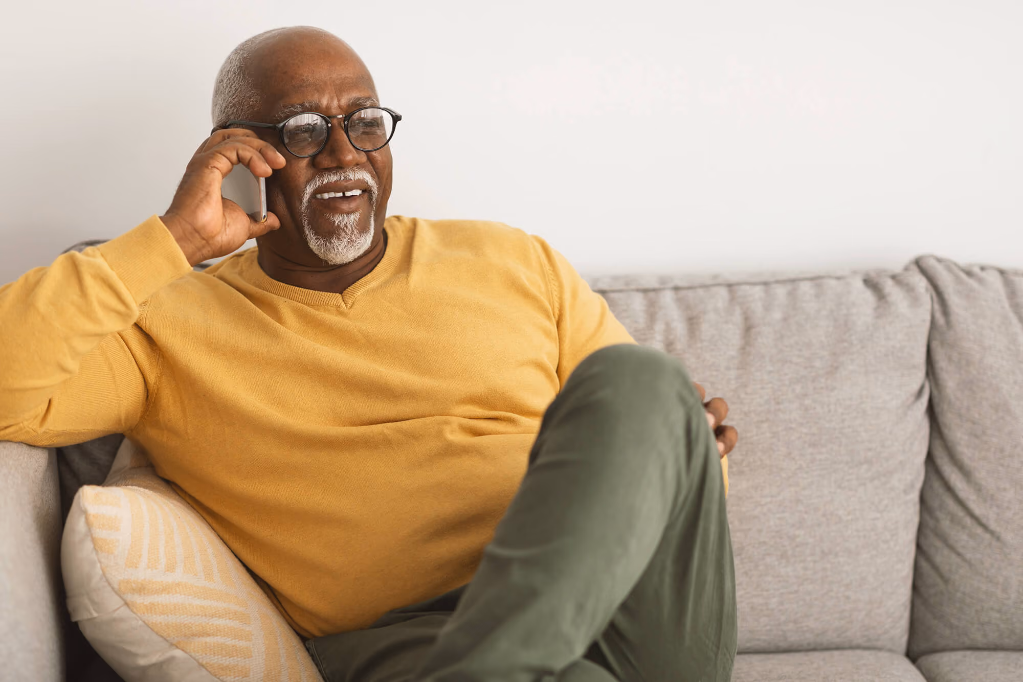 Older man with glasses sitting on a couch, smiling while talking on a phone.