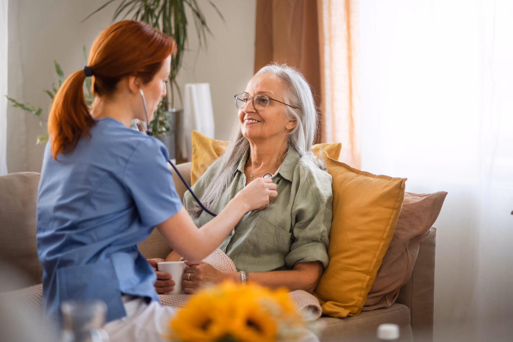 Healthcare worker using a stethoscope to listen to an older woman’s heart while she sits smiling on a couch at home.