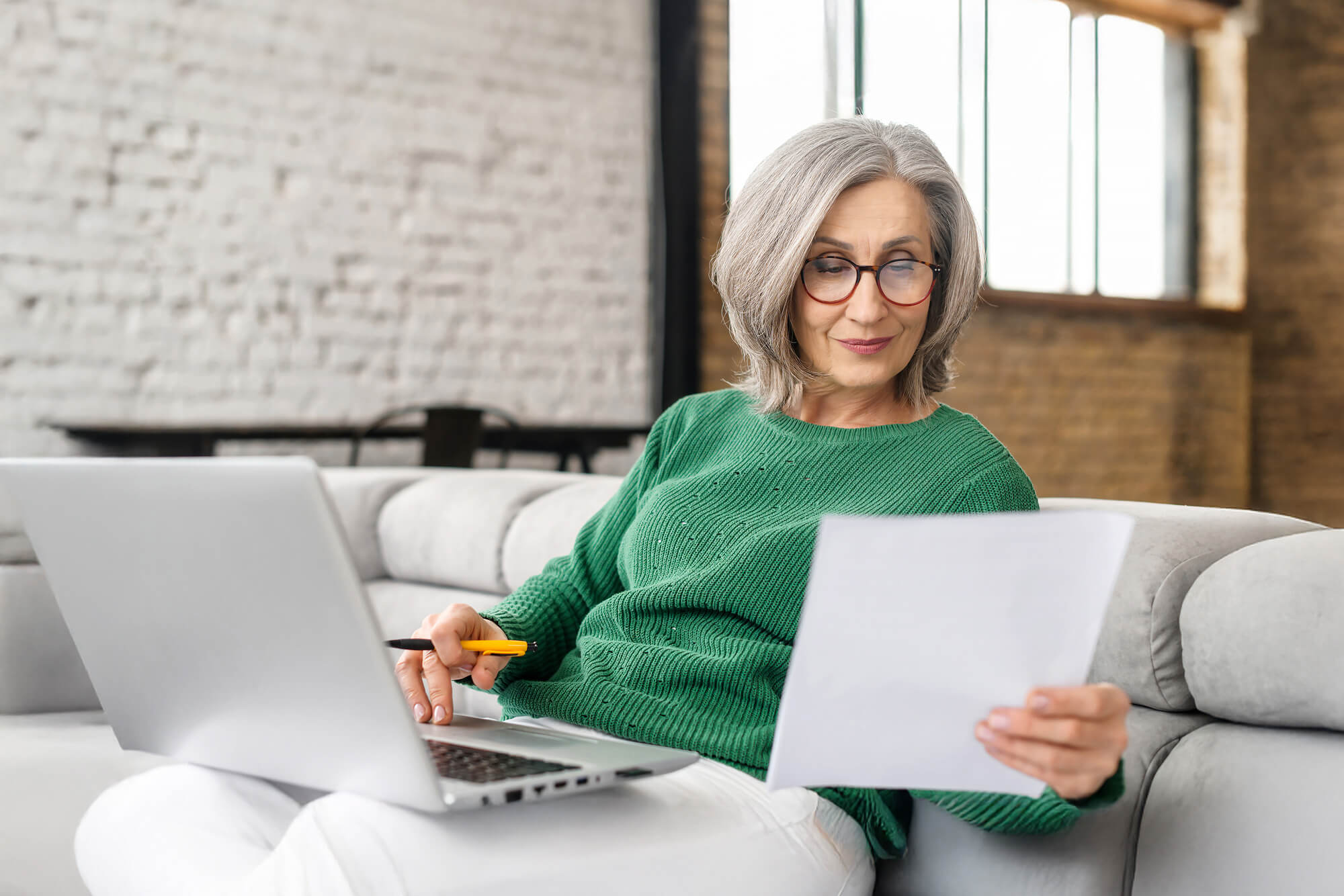 woman reading papers with her laptop on a couch
