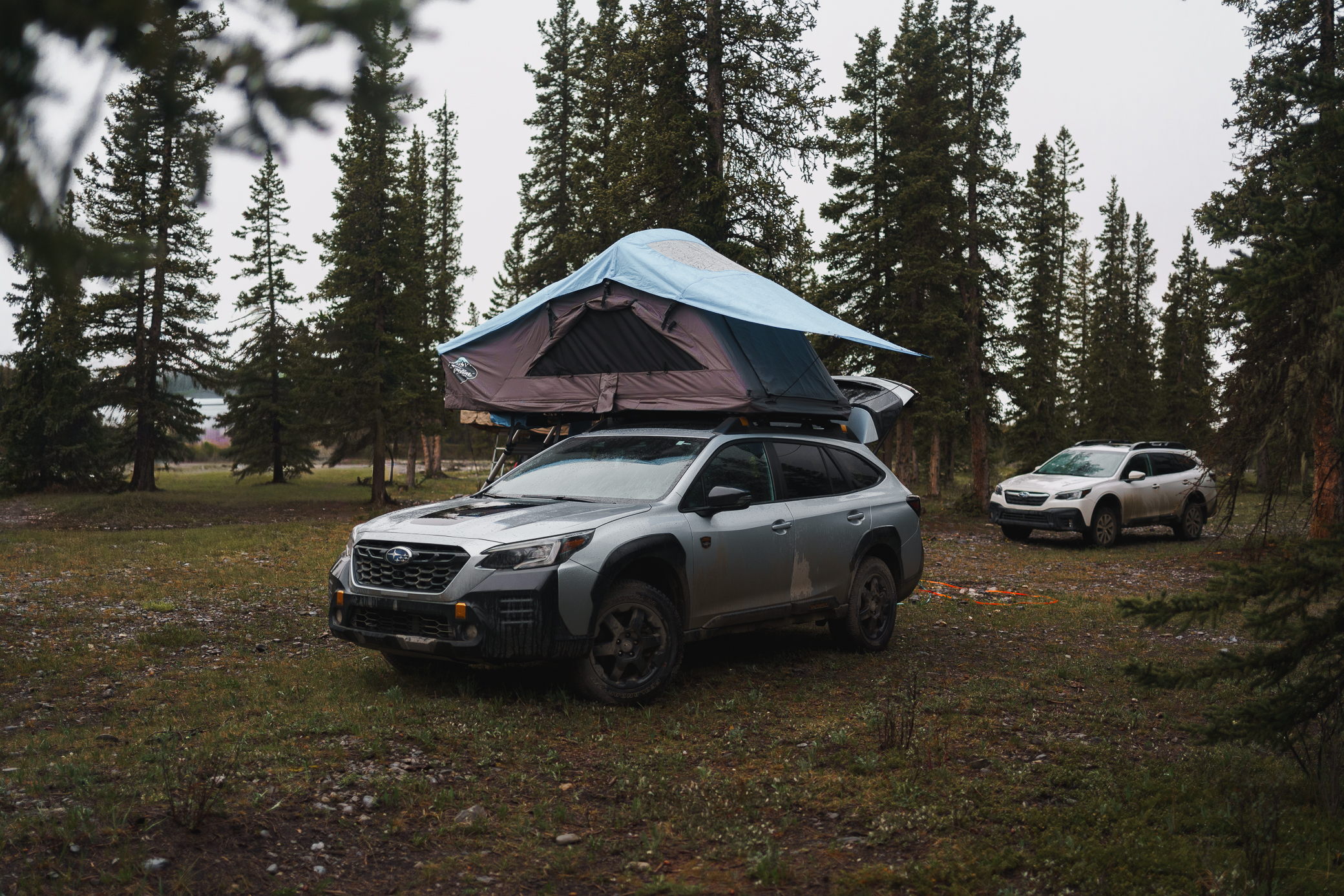 A soft shell rooftop tent open on a lifted Subaru Outback at a forested backcountry campsite in the Canadian Rockies.