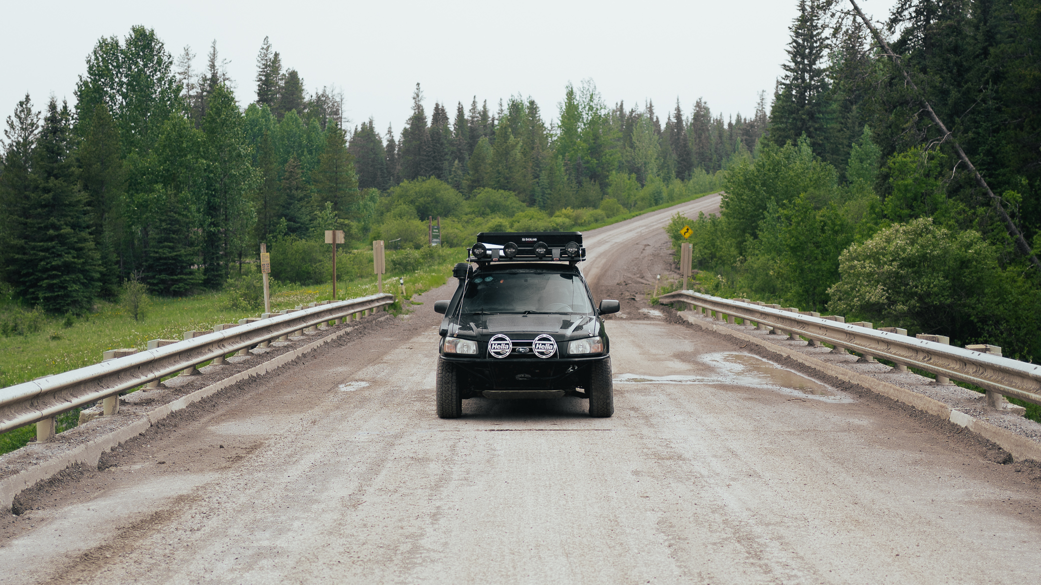 Lifted black Subaru Forester with Go Overland roof rack and Hella driving lights crossing a gravel bridge surrounded by forest