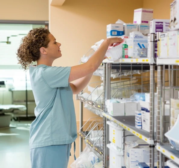 Healthcare worker in blue scrubs organizing medical supplies on metal shelves in a hospital supply room.