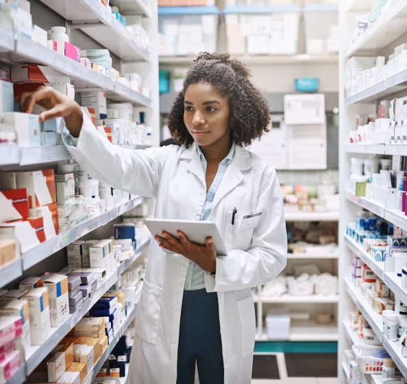 Pharmacist in a white coat holding a tablet and selecting medication from shelves in a pharmacy.