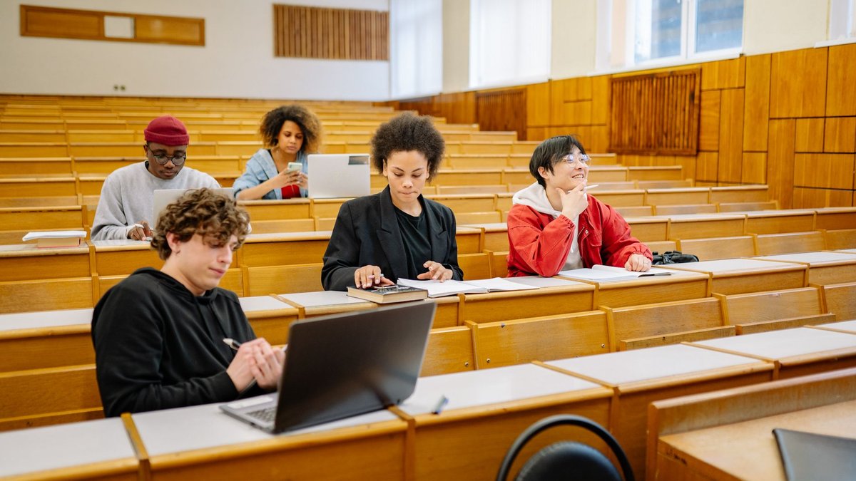 Mehrere Studierende sitzen auf Holzbänken in einem Hörsaal und schreiben mit oder nutzen Laptops