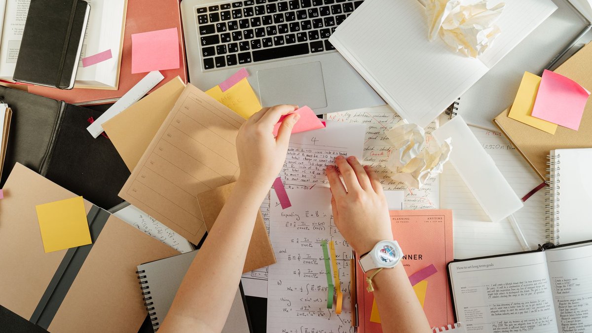 Draufsicht auf einen überfüllten Schreibtisch mit Notizbüchern, Klebezetteln und Laptop, zwei Hände sortieren gleichzeitig mehrere Zettel