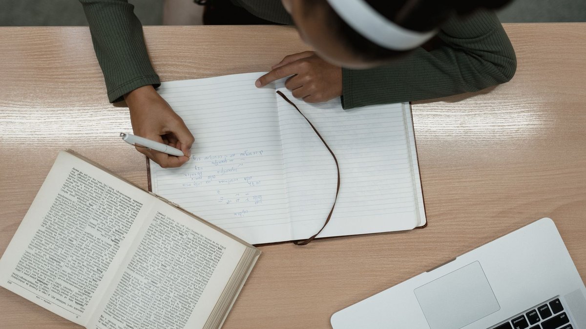 Draufsicht auf einen Holztisch, eine Person schreibt handschriftliche Notizen neben einem aufgeschlagenen Buch und einem Laptop