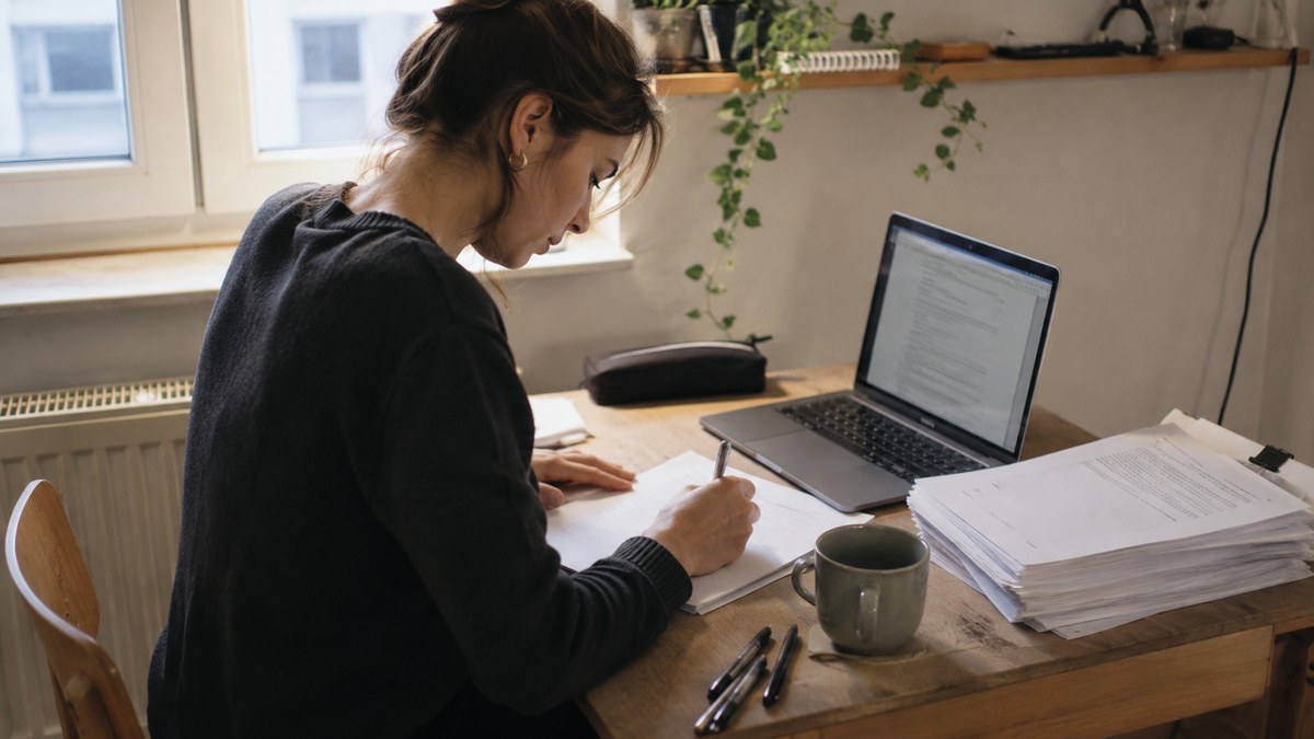 Studentin arbeitet am Schreibtisch am Fenster neben Laptop und einem Stapel Papier