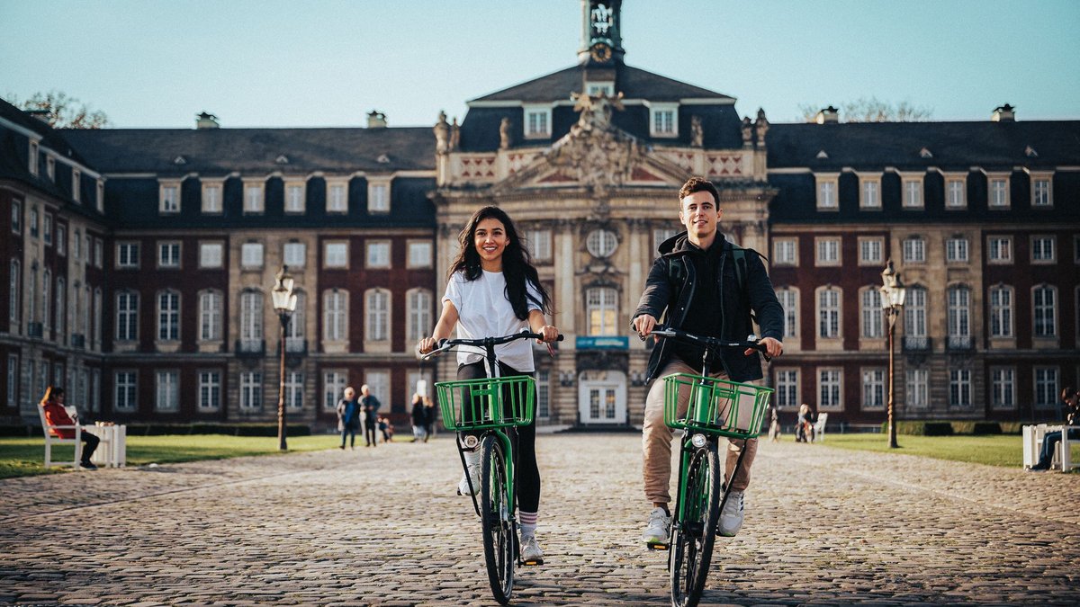 Zwei Studierende fahren mit dem Rad über den Schlossplatz vor dem Schloss der Universität Münster