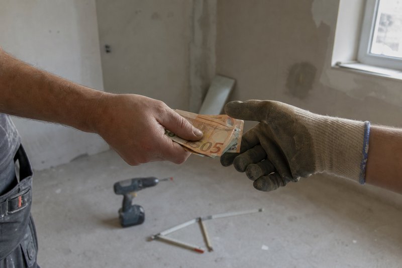 Eine Hand reicht einer behandschuhten Hand Geldscheine in einem unverputzten Rohbau, am Boden liegen Akkuschrauber und Zollstock