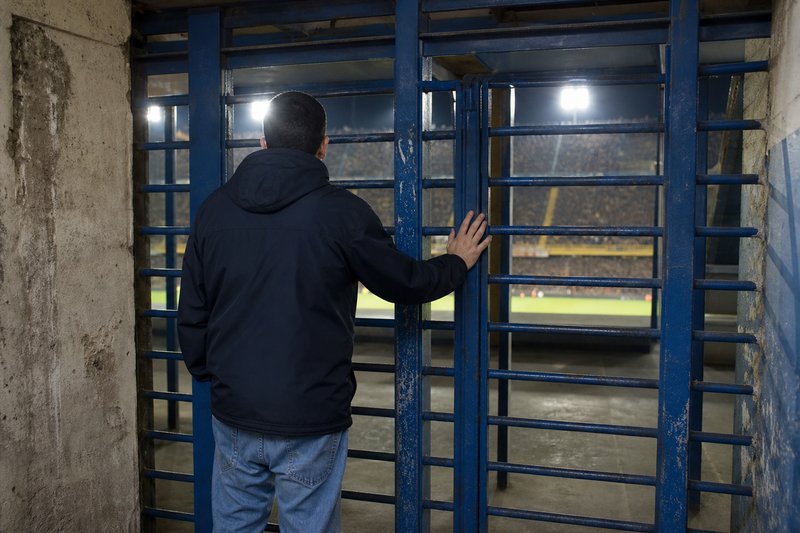 Ein Mann steht von hinten gesehen vor einem geschlossenen Drehkreuz, dahinter läuft unter Flutlicht ein Spiel im vollen Stadion