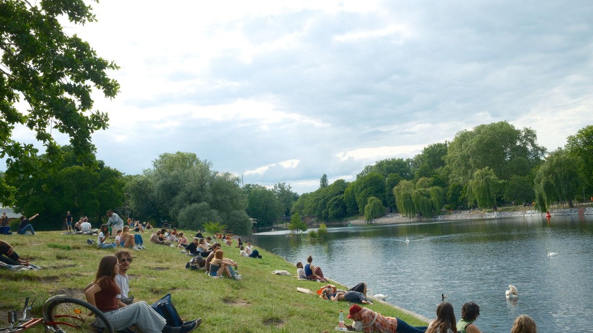 Menschen sitzen an einem Sommertag auf der Wiese am Wasser in einem Berliner Park