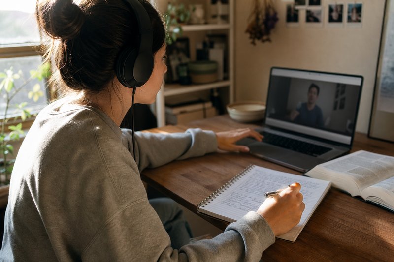 Studentin mit Kopfhörern pausiert ein Vorlesungsvideo am Laptop und schreibt in ihr Notizheft