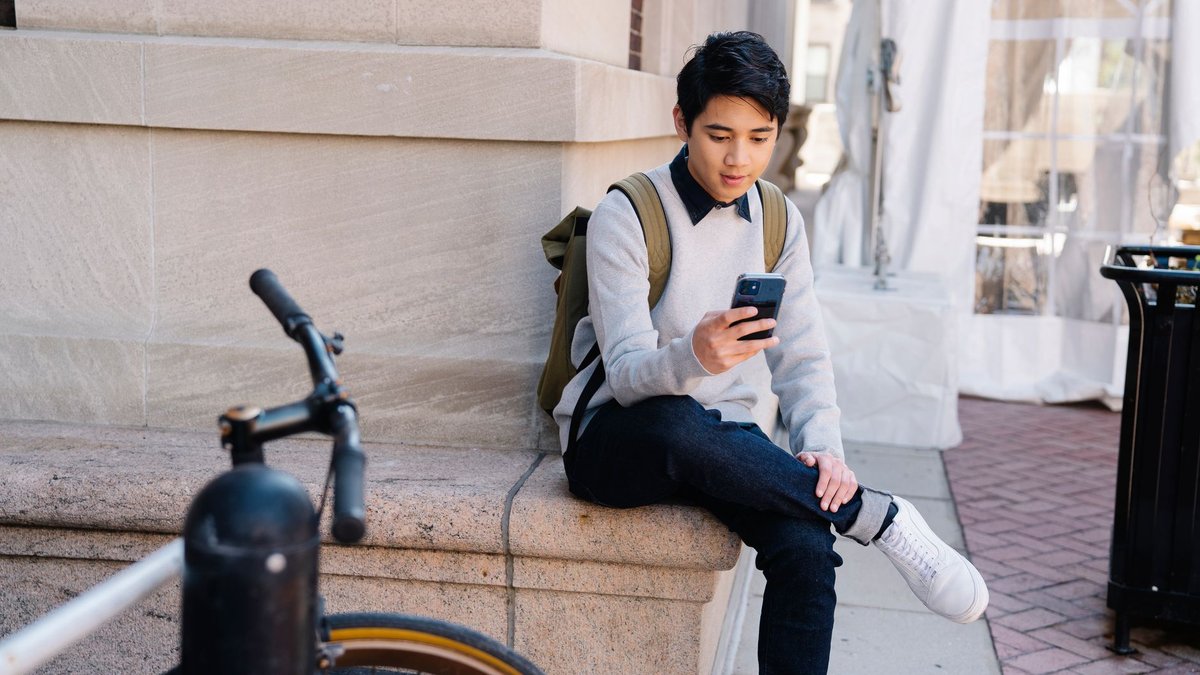 Student mit Rucksack sitzt auf einer Stufe vor einem Unigebäude und schaut auf sein Smartphone