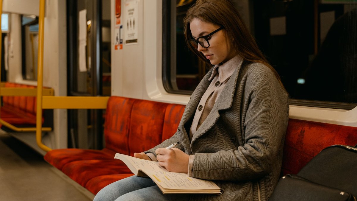 Studentin sitzt in einer U-Bahn und schreibt in ein aufgeschlagenes Notizheft auf ihrem Schoß