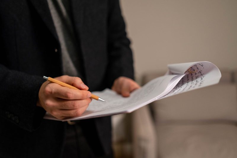 Person im dunklen Sakko hält einen Stapel handschriftlich beschriebener Blätter und geht sie mit einem Bleistift durch