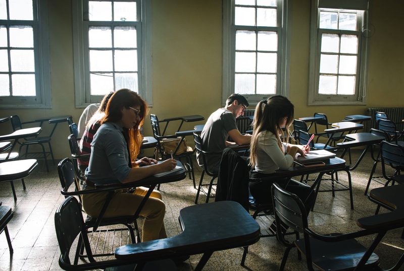 Studierende sitzen in einem sonnendurchfluteten Prüfungsraum an Einzeltischen und schreiben