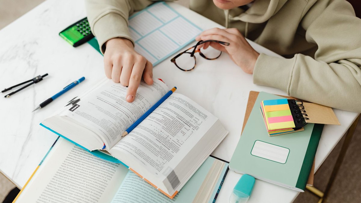 Aufgeschlagenes Lehrbuch mit Taschenrechner, Stiften und Notizheften auf einem hellen Schreibtisch