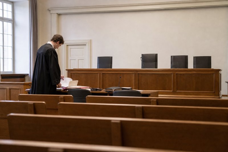 Blick von den leeren Zuschauerbänken auf einen Anwalt in Robe, der am Verteidigertisch seine Unterlagen sortiert