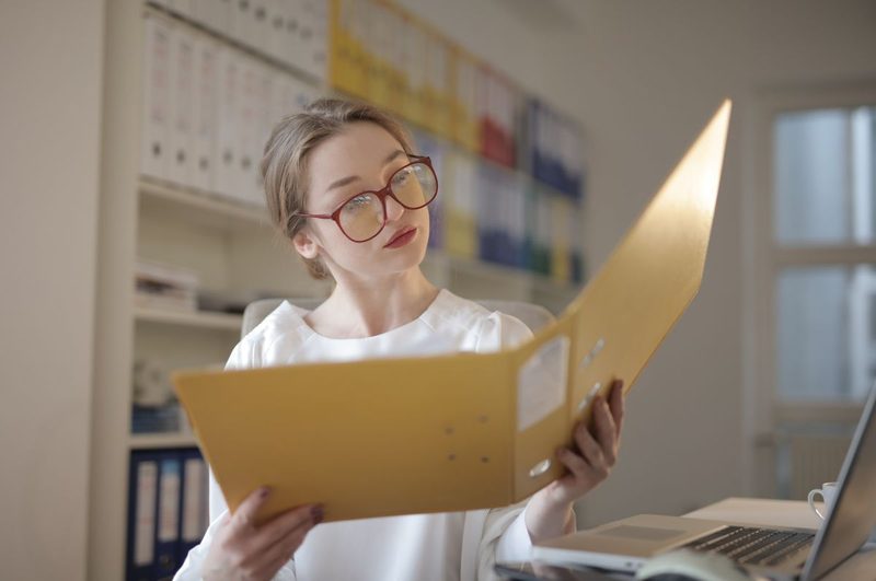 Junge Frau mit Brille liest in einem geöffneten Aktenordner vor einem Regal voller Ordner