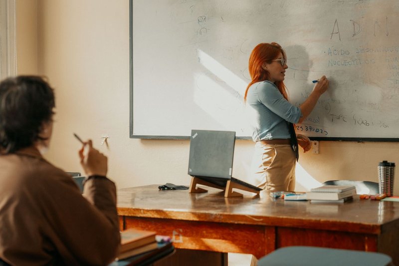 Dozentin schreibt an einem Whiteboard, im Vordergrund folgt eine Studentin mit Notizblock