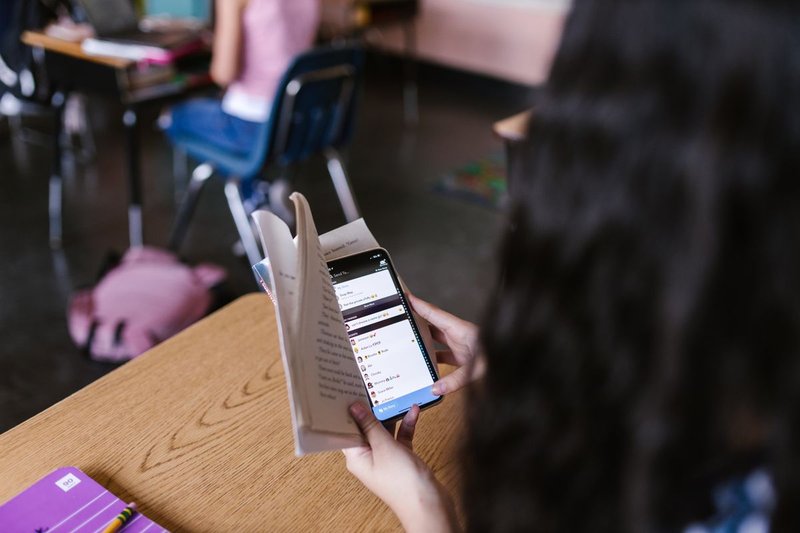 Über die Schulter fotografiert: eine Person hält ein Smartphone vor ein aufgeschlagenes Buch am Lerntisch