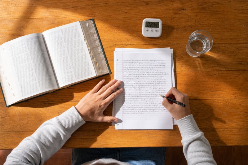 Draufsicht auf einen Tisch: zwei Hände schreiben eine Klausur von Hand, daneben ein aufgeschlagenes Gesetzbuch und eine laufende Küchenuhr