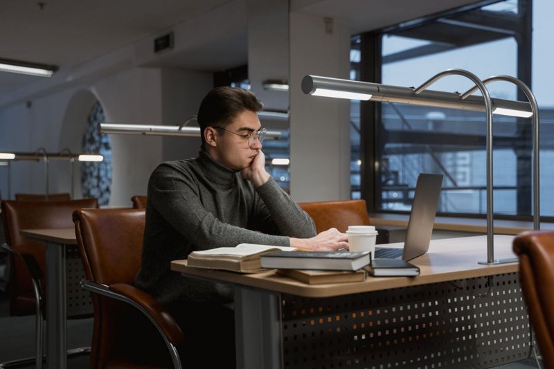 Student sitzt abends allein an einem Bibliothekstisch und recherchiert am Laptop neben aufgeschlagenen Büchern