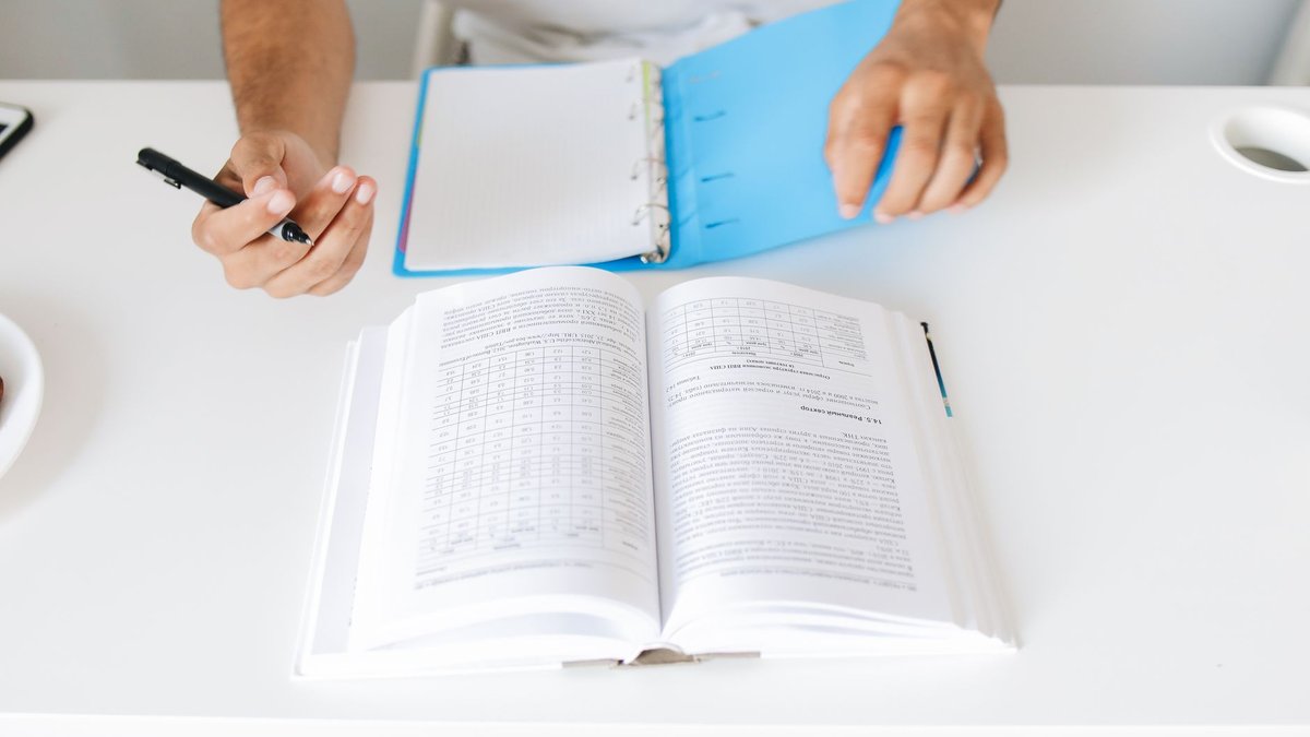 Person blättert mit einem Stift in der Hand in einem aufgeschlagenen dicken Gesetzbuch am Schreibtisch