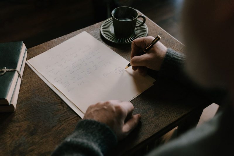 Person schreibt am Abend mit Füller handschriftliche Notizen auf ein Blatt, daneben eine Tasse Kaffee und ein Notizbuch