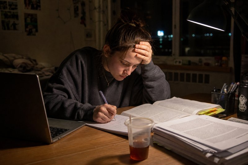 Studentin schreibt spät nachts am Schreibtisch handschriftlich neben einem aufgeschlagenen Kommentar, den Kopf in die Hand gestützt