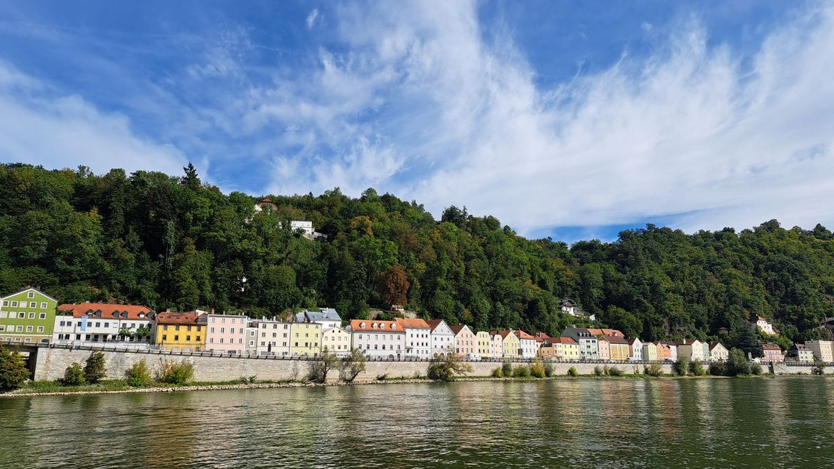 Bunte Häuserzeile am Flussufer in Passau unter blauem Himmel, dahinter bewaldeter Hang