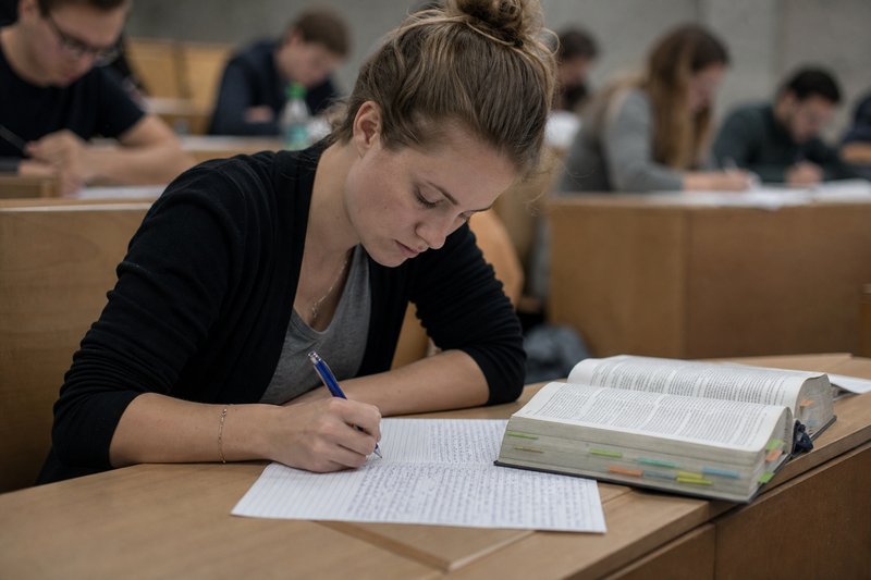 Studentin schreibt im Hörsaal eine juristische Klausur von Hand, daneben ein aufgeschlagener Gesetzestext mit farbigen Reitern