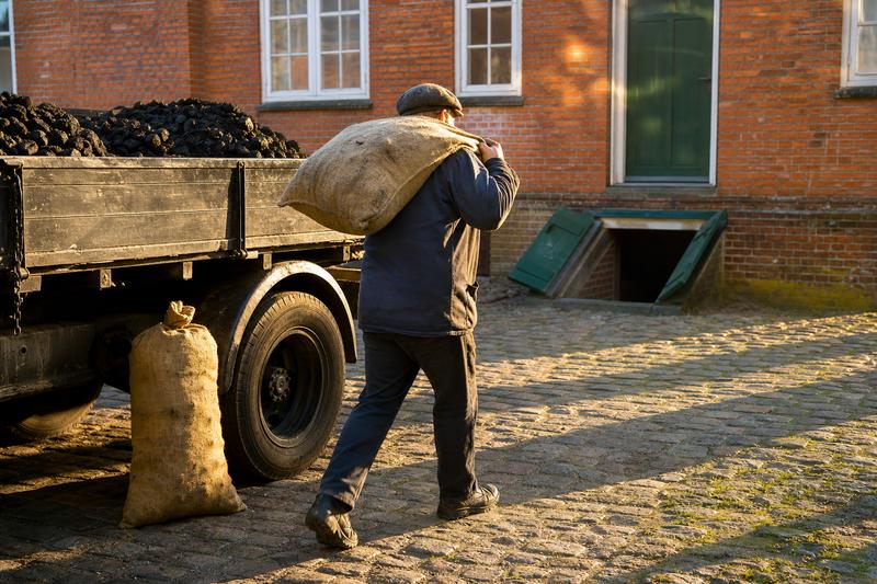 Ein Kohlenhändler trägt einen Jutesack von einer alten Pritsche über einen Kopfsteinpflasterhof zur Kellerluke eines Backsteinhauses