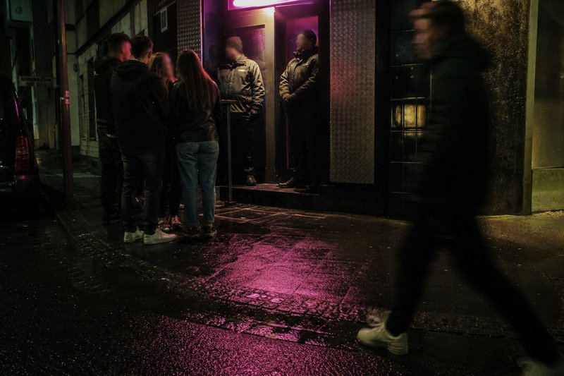 Nächtlicher Eingang eines Clubs, zwei Türsteher an der Tür, eine kleine Gruppe wartet auf dem nassen Gehweg