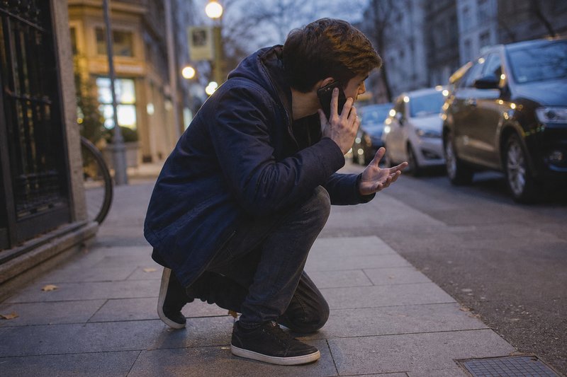 Ein junger Mann kauert auf dem Gehweg und telefoniert aufgeregt mit dem Handy am Ohr