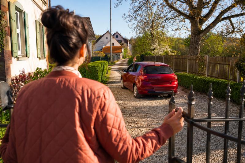 Frau steht am offenen Gartentor ihrer Kiesauffahrt und sieht zu, wie ein rotes Auto über die Auffahrt zum hinteren Weg fährt