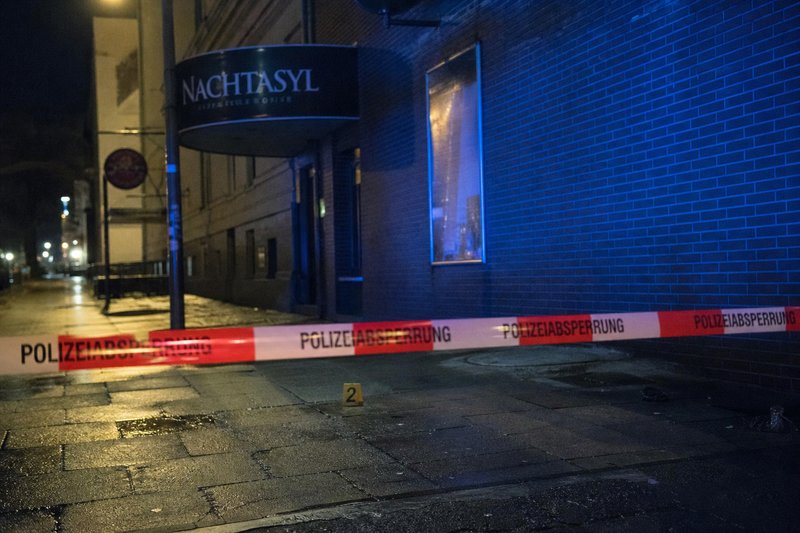 Polizeiabsperrung vor einem geschlossenen Club bei Nacht, dahinter eine gelbe Nummerntafel auf nassem Gehweg im Blaulicht