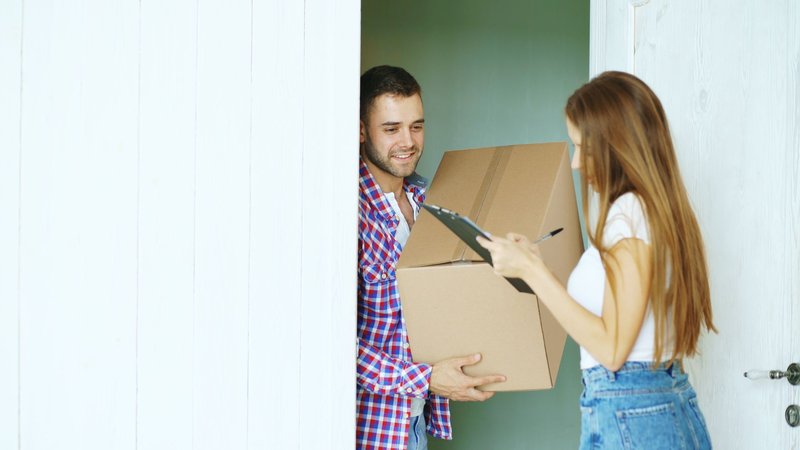 Ein Zusteller übergibt an der Wohnungstür ein Paket, die Empfängerin quittiert auf einem Klemmbrett