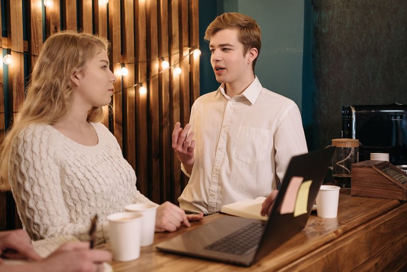 Junger Mann erklärt einer Kommilitonin etwas an einem Cafétisch mit Laptop und Notizbuch, sie hört zu