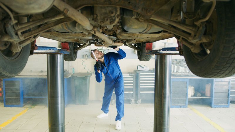 Prüfer in blauer Arbeitskleidung leuchtet unter ein auf der Hebebühne stehendes Auto