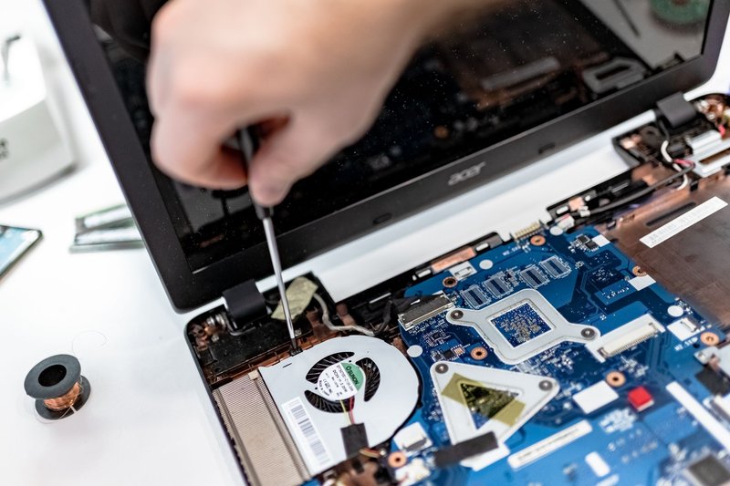 Hand mit Schraubendreher arbeitet an der geöffneten Unterseite eines Laptops mit sichtbarer Hauptplatine und Lüfter