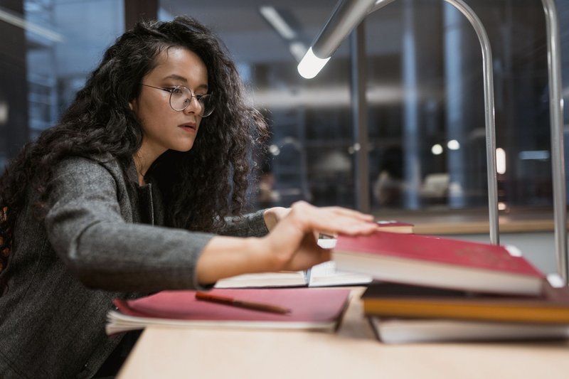 Studentin mit Brille greift an einem Bibliothekstisch unter einer Leselampe nach einem roten Gesetzbuch