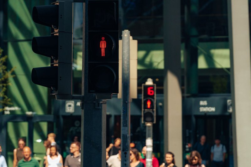 Rote Fußgängerampel im Vordergrund, dahinter Menschen an einer Straßenkreuzung